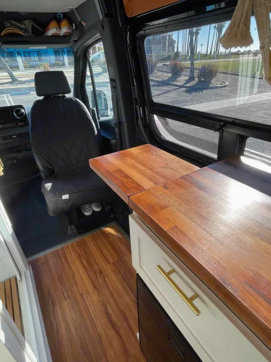 Inside an RV rental: custom wood floor & countertop, driver's seat, and scenic beach view.. Mercedes-Benz Sprinter 2021