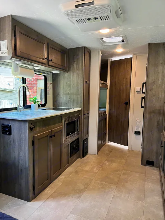 Modern rv rental interior: kitchen with sink, microwave, dark wood cabinets, and light tiled floor. Ceiling AC unit visible.. Coachmen Apex Nano 2021