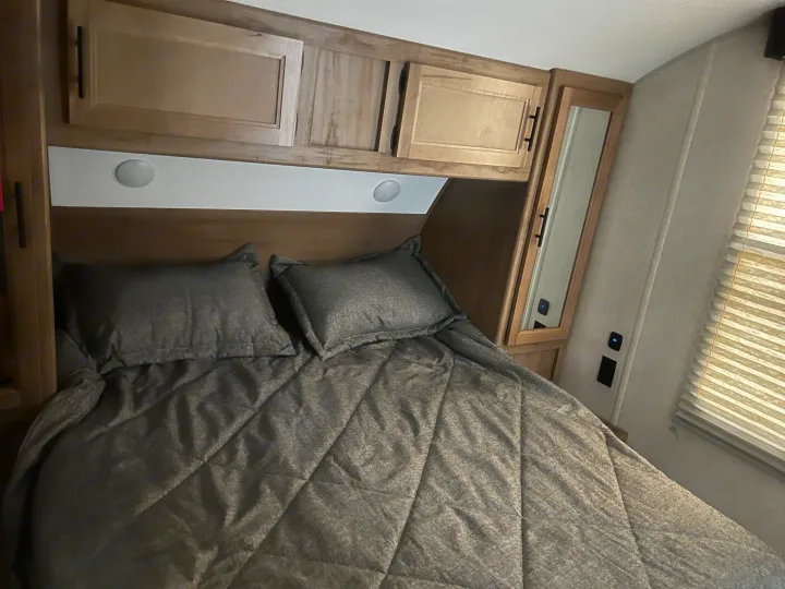 This RV rental bedroom offers a comfortable gray bed, plump pillows, overhead wood cabinets, and convenient bedside lighting.. Forest River 29TE 2023