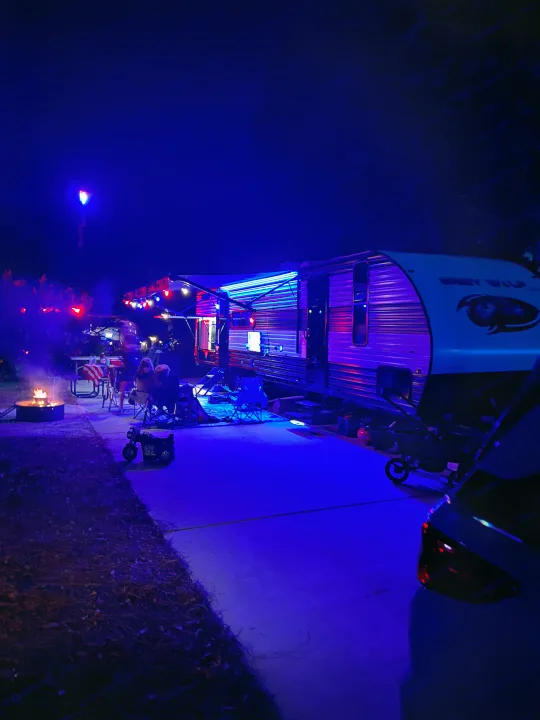 Relaxing night at an RV rental: Grey Wolf illuminated by vibrant blue and purple lights, people gathered by a campfire.. Forest River 29TE 2023