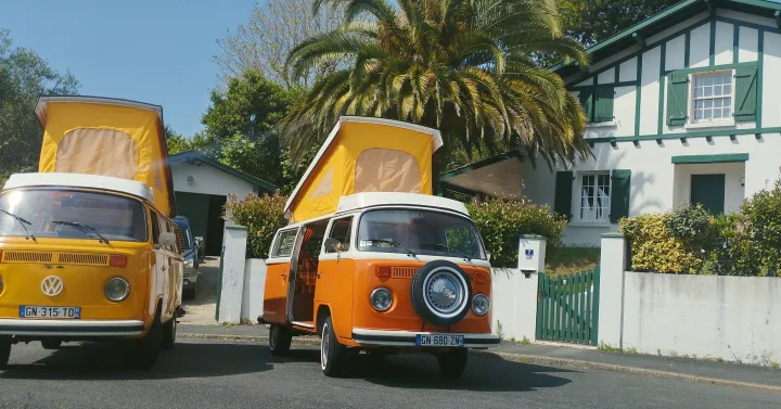 Two vintage RV rental vans, one yellow and one orange, with extended pop-tops, parked on a sunny street beside a white house.. Volkswagen T2 Westfalia 1978