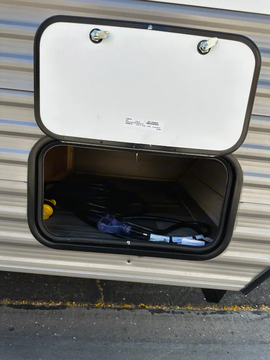 An RV rental's exterior storage compartment is open, revealing a coiled black power cord with a yellow adapter inside.. Dutchmen Coleman Lantern LT 2023