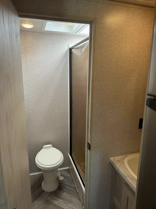 Clean, compact RV rental bathroom showing toilet, roll-away shower door, and sink area.. East to West Della Terra 2023