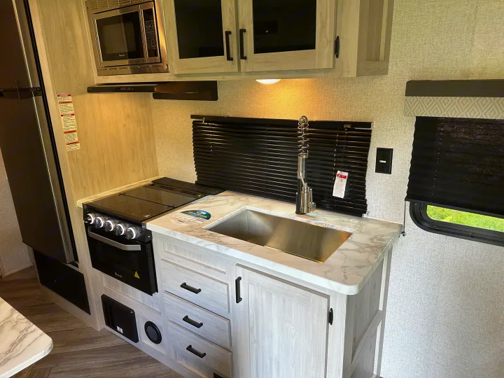 A fully-equipped, modern RV rental kitchen with a microwave, stove, sink, and fridge, showcasing light cabinets and marble counters.. East to West Della Terra 2023