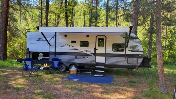 Enjoy nature with an RV rental! This travel trailer is parked in a sunny forest campsite with outdoor seating and kitchen.. Jayco Jay Flight 2023
