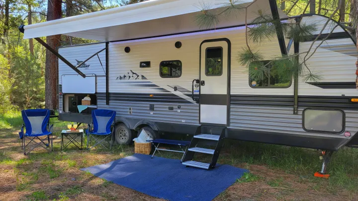 Relaxing outdoors with an RV rental. This travel trailer features an extended awning, chairs, and mat at a serene campsite.. Jayco Jay Flight 2023