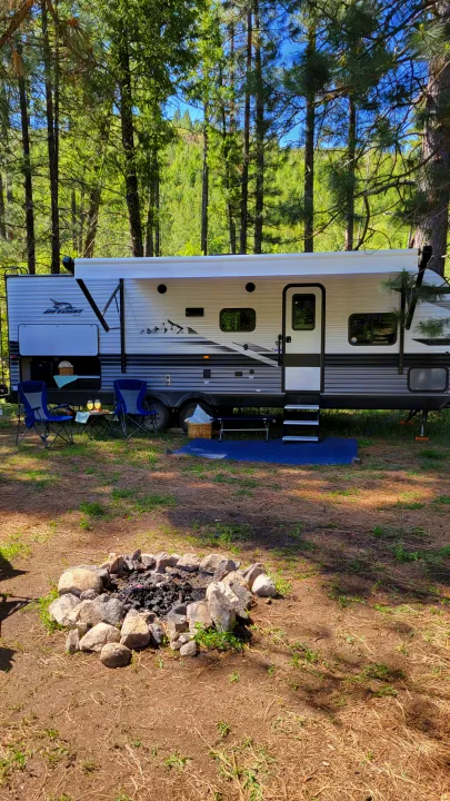 Enjoy an RV rental adventure! This travel trailer is parked in a sunny forest campground, with chairs and a fire pit ready for relaxation.. Jayco Jay Flight 2023