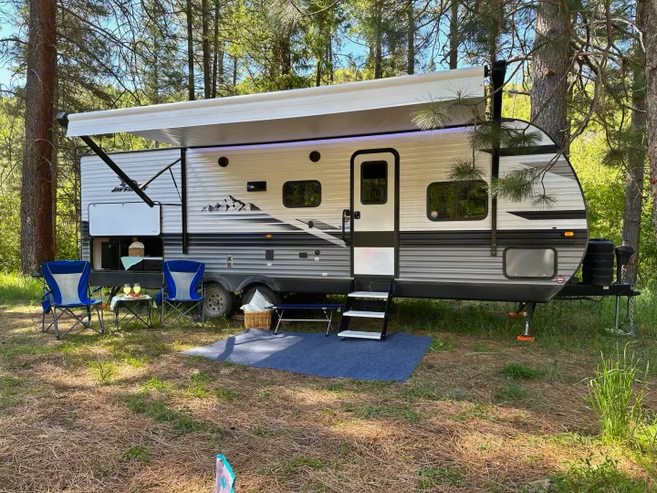 Enjoying the outdoors with an rv rental. A modern travel trailer is set up with chairs, a table, and mat in a sunny, wooded campsite.. Jayco Jay Flight 2023