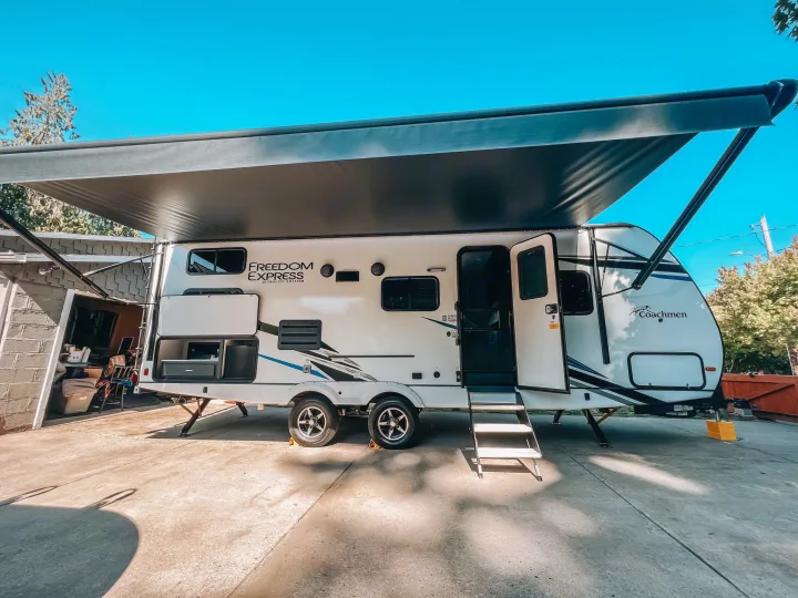 A white Coachmen Freedom Express travel trailer, awning extended & steps down, on a driveway. Great rv rental for adventures.. Coachmen 257BHS 2022