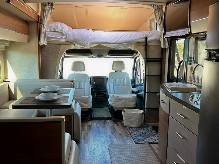 Bright RV rental interior showing a dinette, galley kitchen, front cab seats, and an accessible overhead bunk bed.. Winnebago View 2017