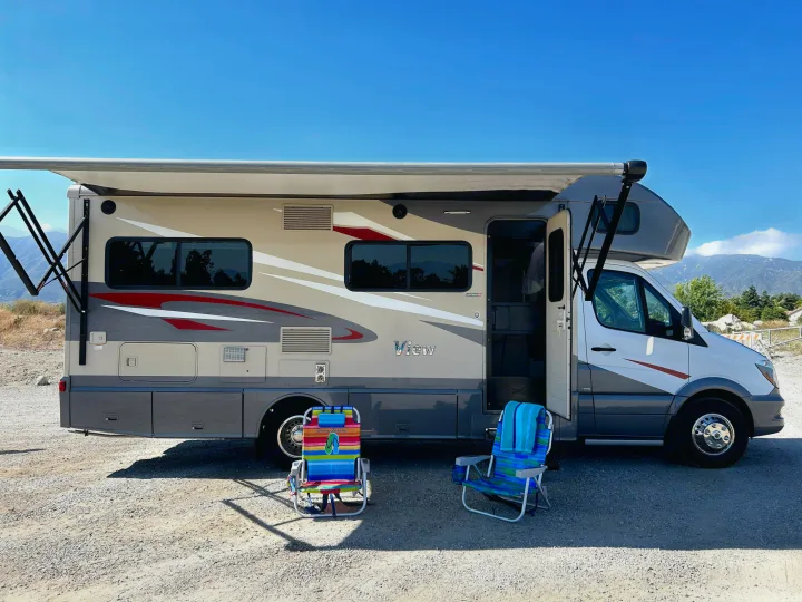 Scenic rv rental perfect for outdoor adventures! Beige RV with awning & colorful chairs parked against a mountain backdrop.. Winnebago View 2017