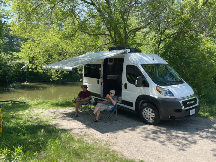 Two people relax in chairs by a river, next to their white rv rental camper van with awning extended, under sunny trees.. Thor Motor Coach Scope 2022