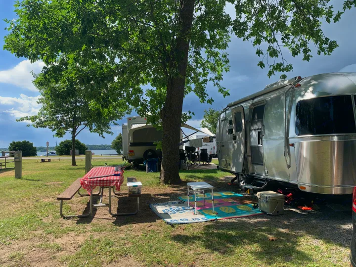 A serene rv rental campsite by a lake with a shiny Airstream, picnic table, and mats. Enjoy shaded outdoor RV camping.. Airstream Caraval 2021