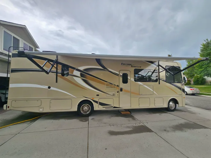 Large tan motorhome with its awning extended and windows open, parked on a driveway, perfect for your RV rental travel.. Thor Freedom Traveler 2020
