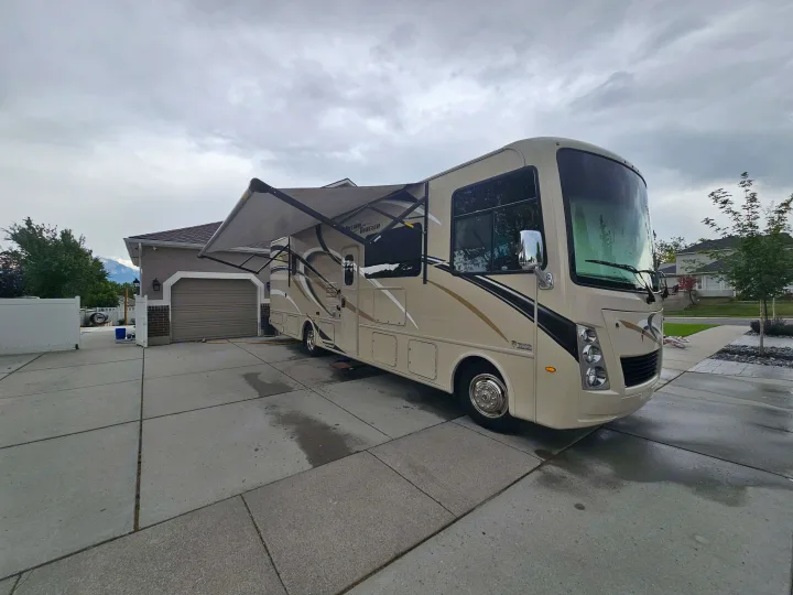 Spacious tan RV rental parked on a wet driveway with awning out, ready for travel adventures under cloudy skies.. Thor Freedom Traveler 2020