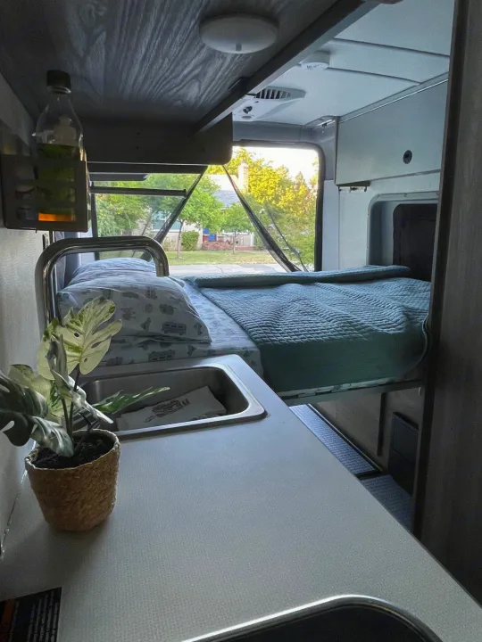Cozy RV rental interior with a compact sink/counter, bed, and window view of trees. Perfect for adventure travel.. Winnebago Solis 2023