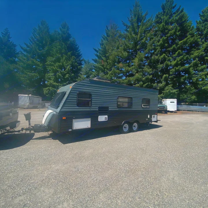 Dark gray travel trailer/RV rental on a sunny gravel lot, hitched to a truck, with trees in the background.. Fleetwood Prowler 1996