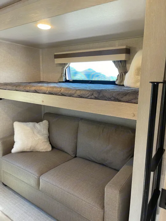 Cozy RV rental interior: upper bunk with a grey blanket, lower sofa with a white pillow, and a window framing mountain views.. Forest River Rockwood Signature 2023