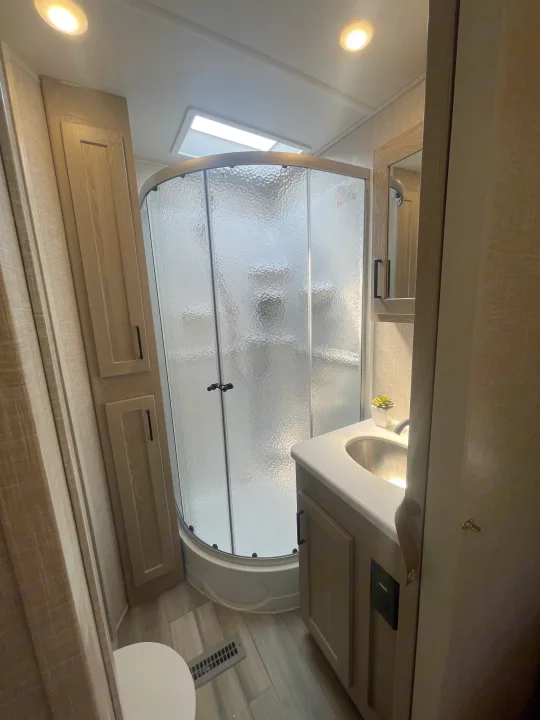 An RV rental bathroom featuring a compact design with a curved glass shower, sink vanity, and toilet.. Forest River Rockwood Signature 2023