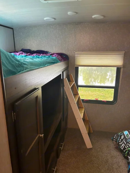 Cozy RV rental interior: a top bunk bed with colorful bedding, a wooden ladder, and a window revealing an outdoor scene.. Heartland RVs Mallard 2020