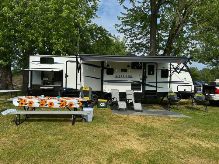 Enjoy outdoor living with this RV rental! A white travel trailer is set up at a sunny campsite with a picnic table and lounge chairs.. Heartland RVs Mallard 2020