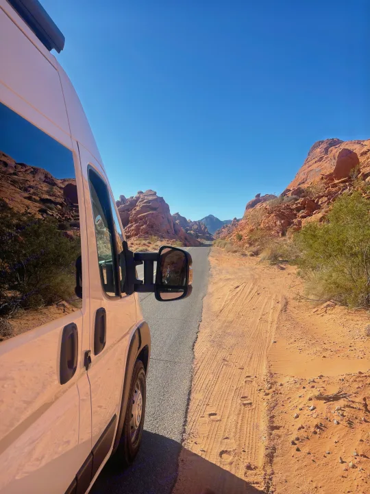 White RV rental on a desert road, flanked by towering red rock formations under a clear blue sky.. Dodge Ram Promaster 3500 C/V HR 159 EXT 2023