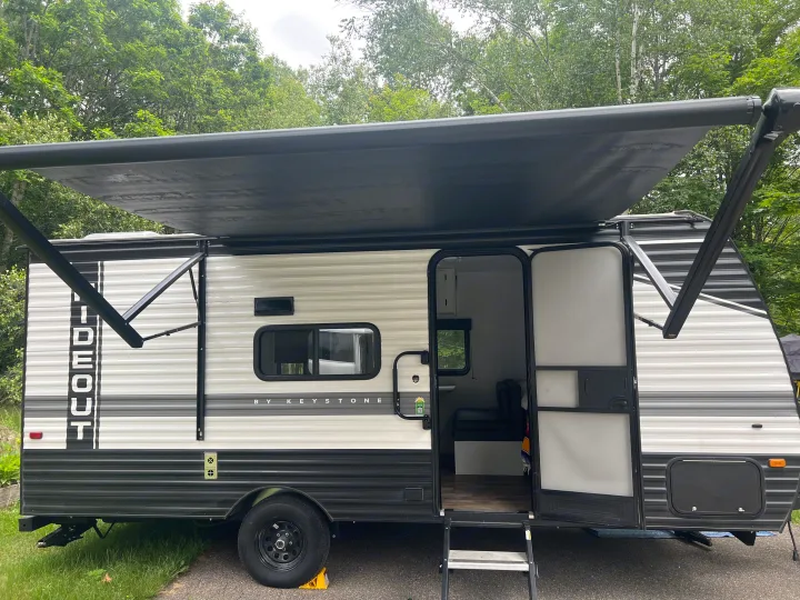 A white and black Keystone travel trailer, an inviting RV rental, parked at a green campsite with awning extended.. Keystone RV Hideout 2023