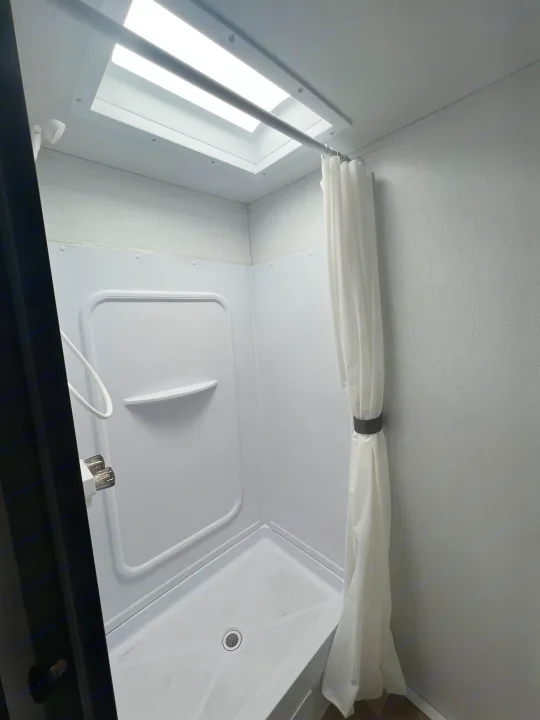 Inside a modern RV rental shower, featuring a bright skylight, compact white stall, and curtain.. Keystone RV Hideout 2023