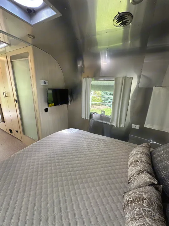 Comfortable RV rental bedroom with a sleek Airstream interior, showing a bed, TV, and a window with greenery outside.. Airstream Flying Cloud 2022