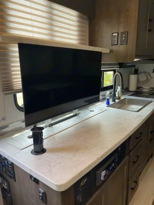 Interior of an RV rental: modern kitchen with a pop-up TV, sink, and white marble-look counter. Features integrated stereo.. Coachmen Leprechaun 2023