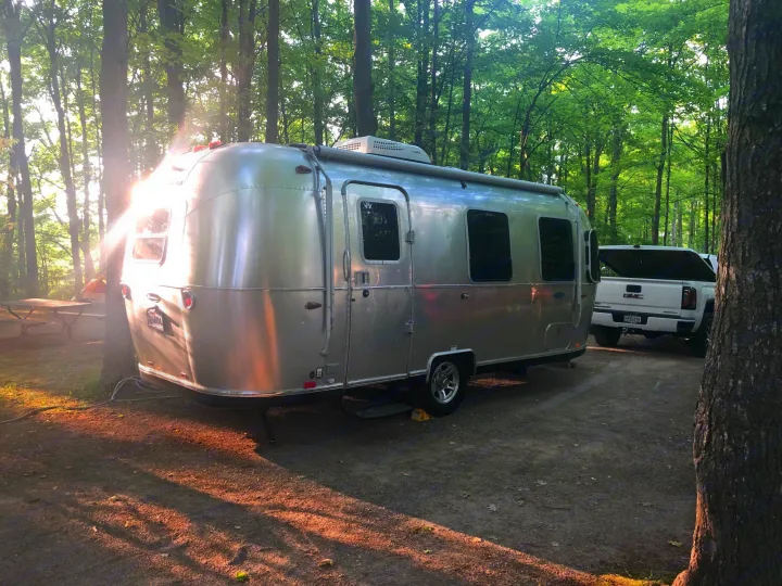 A silver Airstream RV rental shines at a serene, sun-dappled campsite with a GMC truck amidst tall trees.. Airstream Sport 2019