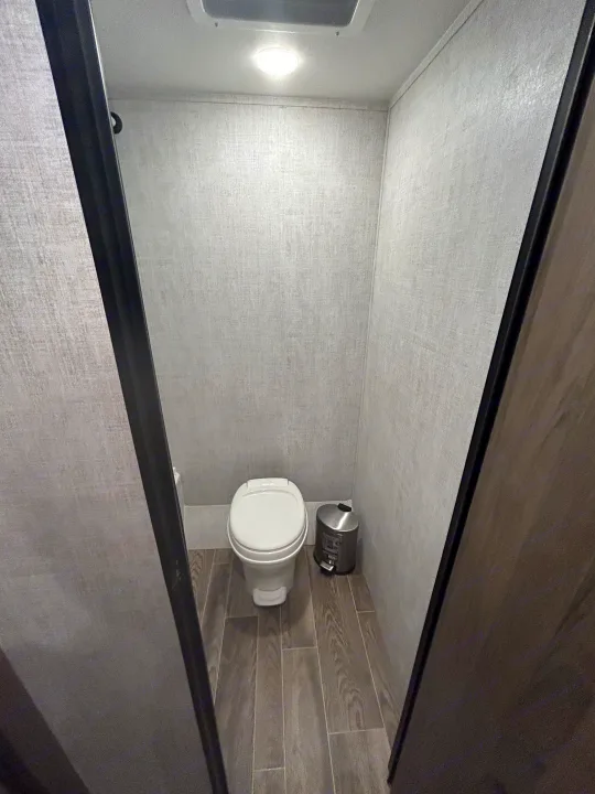 Compact RV rental bathroom features a white toilet, gray wood-look floor, and light walls, visible through a doorway.. Gulf Stream Ameri-Lite Super Lite 2021