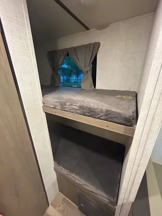 Interior of an RV rental showing two cozy bunk beds with plush grey bedding, and a window with curtains offering an outside view.. Gulf Stream Ameri-Lite Super Lite 2021