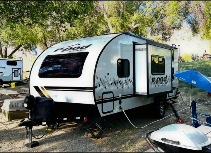 A sleek r-pod travel trailer, a popular RV rental, is set up at a shady campsite with outdoor gear like a grill and umbrella.. Forest River R POD 193  H.R. EDITION 2022