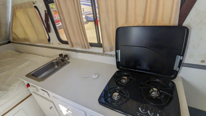 This RV rental interior features a compact kitchen with a sink and gas stove, plus a cozy bed area and windows with curtains.. Chalet Chalet Popup Trailer 2006