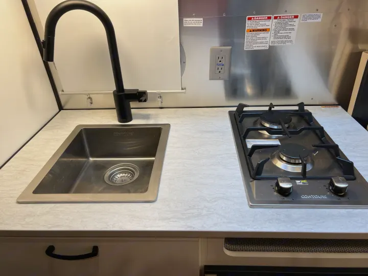 Compact RV rental kitchenette featuring a stainless steel sink with a black faucet and a two-burner gas stove on a light countertop.. Airstream Caravel 2020