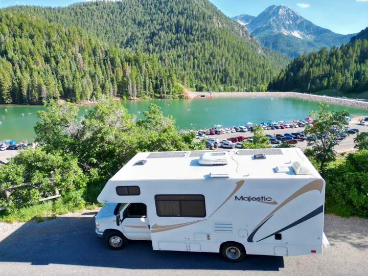 Scenic rv rental view: A white RV overlooking a green lake with boats, surrounded by dense forests and distant snowy mountains.. Thor Majestic 2014