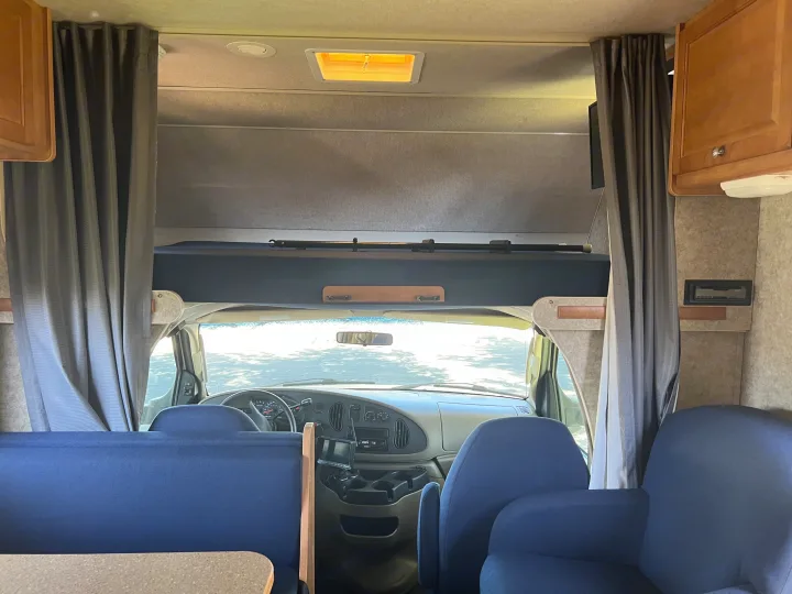 RV rental interior: View from the living area toward the front cab, with blue seating, dashboard, and overhead bunk.. Winnebago Chalet 2010