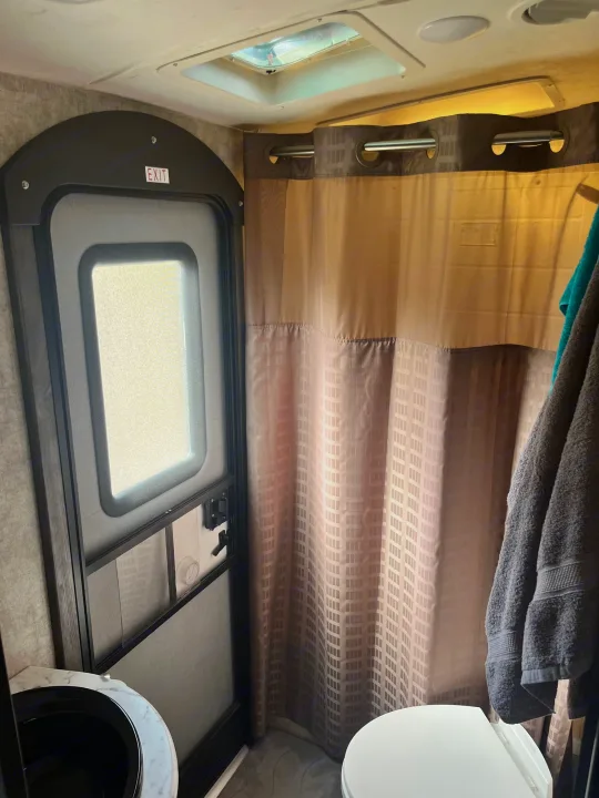 RV rental bathroom: exit door with sign, compact sink, toilet, and patterned shower curtain. Overhead skylight illuminates the space.. Highland Ridge RV Light 2017