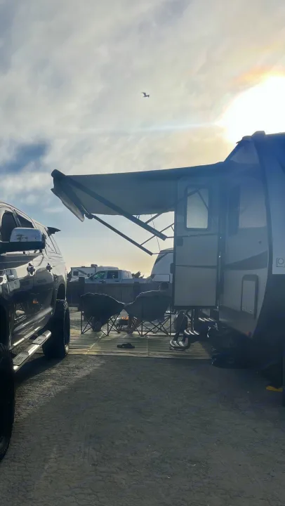 Perfect RV rental setup! Two people relax under the awning of a travel trailer, with a truck parked beside them at a sunny campsite.. Highland Ridge RV Light 2017