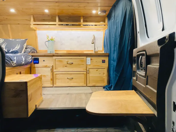 Cozy, custom-built wooden camper van interior: bed with patterned bedding, kitchen with sink/backsplash, and a rear pull-out table, great for RV rental.. Gmc Savanna 2013