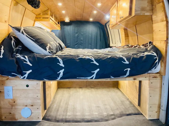 RV rental: a cozy van interior with a comfortable bed, custom wooden walls, overhead cabinets, and under-bed storage with an outlet.. Gmc Savanna 2013