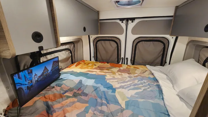 Inside a cozy RV rental: a comfy bed with colorful comforter, TV, and practical overhead storage for travel.. Winnebago Solis 2023