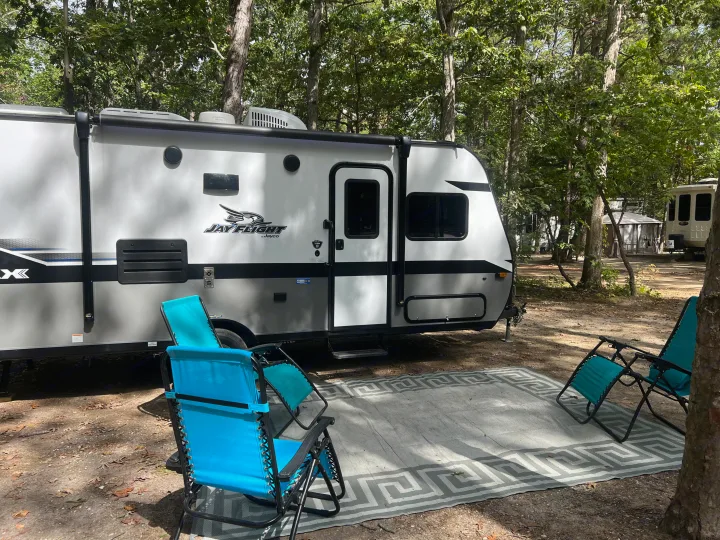 Enjoying the outdoors with an RV rental. A modern travel trailer is set up with blue chairs and a rug at a wooded campsite.. Jayco Jay Flight 2022