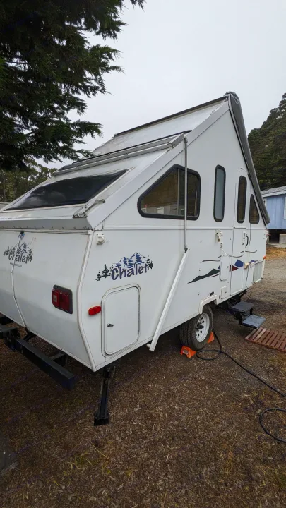 White A-frame Chalet camper, ideal for an RV rental adventure, parked under trees on a cloudy day.. Chalet Chalet Popup Trailer 2006