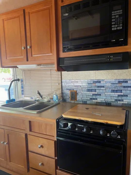 Well-equipped RV rental kitchen featuring wood cabinets, microwave, Magic Chef stove, double sink, and tiled backsplash.. Winnebago Chalet 2010