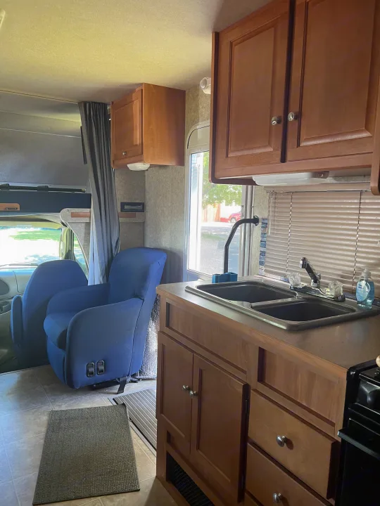 RV rental interior showing a kitchen with wood cabinets & double sink, a comfy blue armchair, and front bunk area.. Winnebago Chalet 2010
