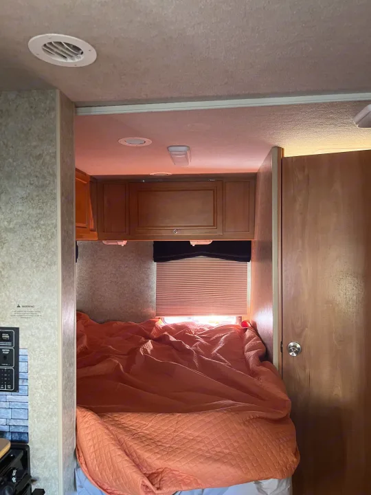 Cozy RV rental bedroom featuring a warm orange quilted comforter, overhead wooden cabinets, and a window with blinds.. Winnebago Chalet 2010
