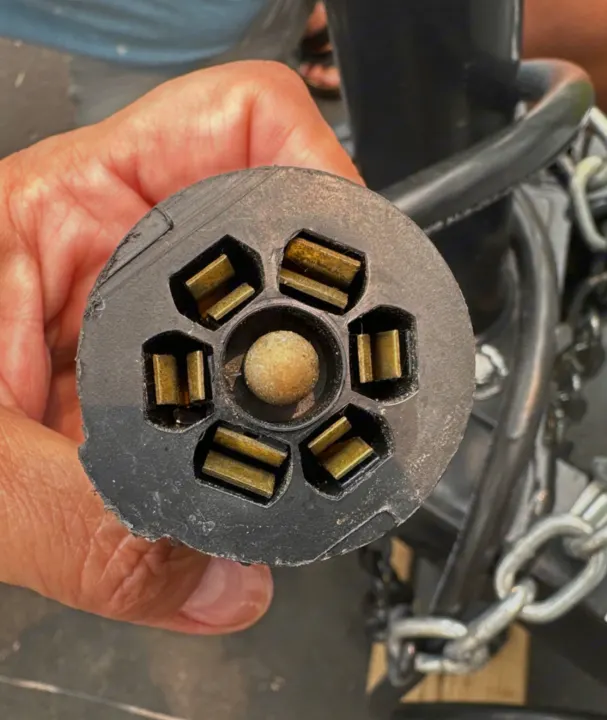 Inspecting an RV rental's 7-way trailer electrical plug. A hand holds the black, round connector with six brass pins and a central one.. Braxton Creek Bushwhacker Plus 2022