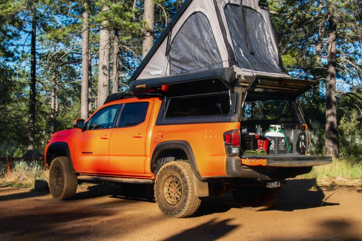 Orange Tacoma overlander with pop-up tent & open bed storage in a sunny forest. Perfect RV rental alternative for adventure.. Toyota Tacoma TRD Pro 2023